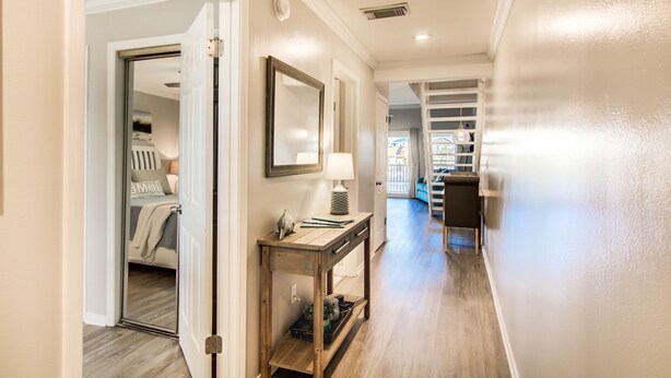 Un pasillo con una consola de madera, un espejo y una lámpara. Hay una puerta que da a un dormitorio con una cama y una mesita de noche.