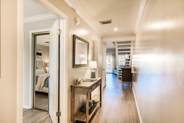 A hallway with a wooden console table, a mirror, and a lamp. There is a door leading to a bedroom with a bed and a nightstand. - Miramar's 2 Min Walk to the Beach with Private access! (Destin)