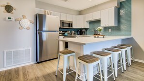 Una cocina moderna con armarios blancos, un refrigerador de acero inoxidable, un microondas y un salpicadero color turquesa. Hay cuatro taburetes de madera alrededor de la isla.