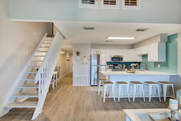 Una cocina moderna con una isla central, taburetes blancos y una escalera que sube a un nivel superior.