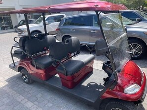 Une voiturette de golf rouge, avec deux sièges noirs et un toit, est garée sur un parking avec d’autres véhicules.