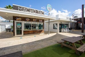 Ein Food Truck mit dem Namen 'MACHO TACO', einem Schild mit der Aufschrift 'LIVE WELL', Sitzgelegenheiten im Freien und einem Schild für 'Wein' im Hintergrund.