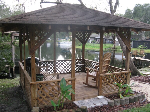 A wooden gazebo with a lattice floor, surrounded by a pond and benches.
