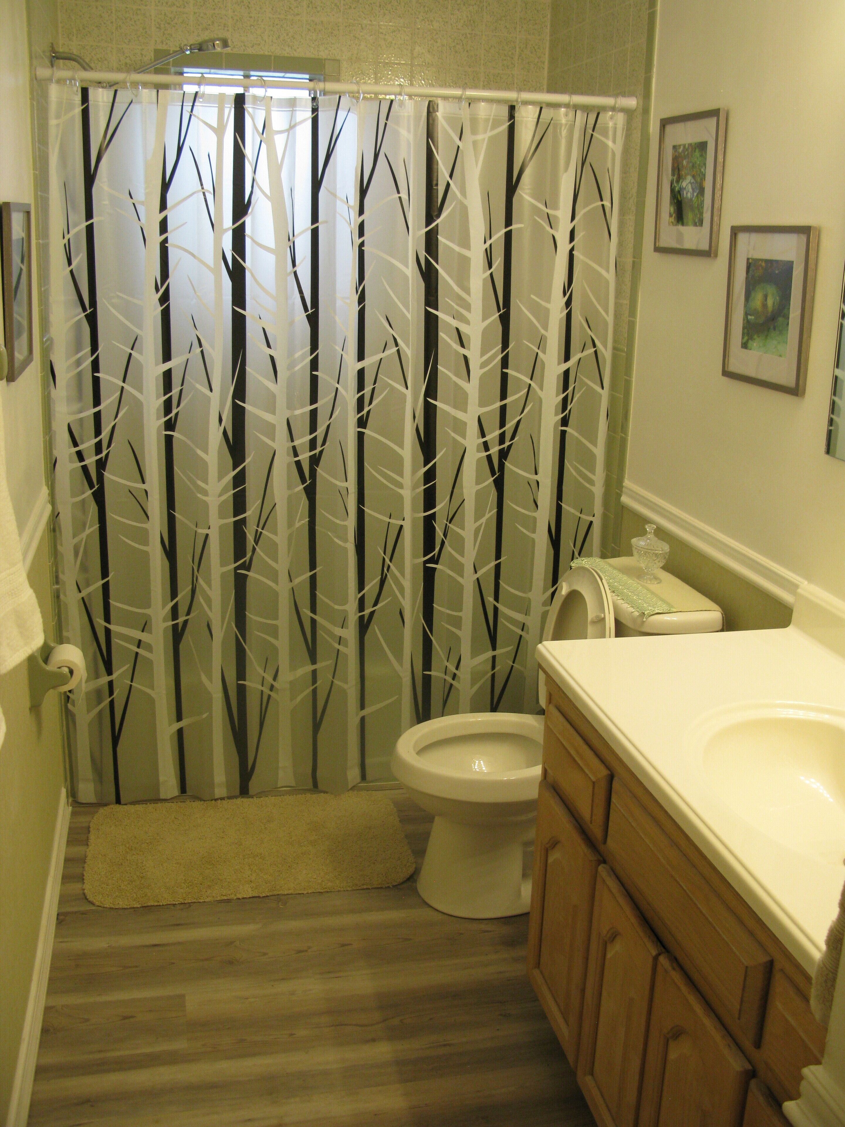 A bathroom with a shower curtain featuring a tree pattern, a toilet, wooden cabinets, and a small rug on the floor.