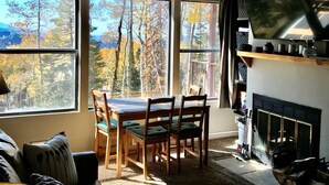 Un salon chaleureux avec une cheminée, une table à manger avec des chaises et de grandes fenêtres donnant sur la forêt.