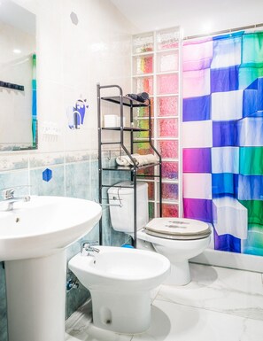 A bathroom with a white sink, toilet, and bidet, a metal shelf unit, and a colorful glass shower curtain. - El Rincón de Maria y Pedro (Torremolinos)