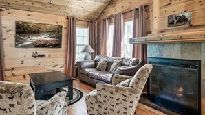 A cozy cabin interior with a fireplace, wooden walls, a leather sofa, patterned chairs, and a painting of a river. - 10 Mins to Downtown Gatlinburg~HotTub~Rockytop (Gatlinburg)