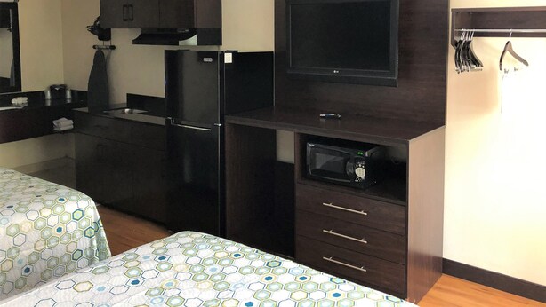 A compact hotel room with a kitchenette, a flat-screen TV, a microwave, and a bed with a patterned bedspread. - HomeTowne Studios by Red Roof Bordentown - McGuire AFB (Bordentown)