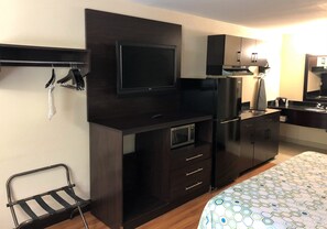 Een compacte hotelkamer met een flatscreen televisie boven een donker houten dressoir, een magnetron, een koelkast, en een klein zithoekje met een stoel.