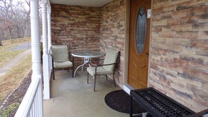 A covered porch with a brick wall, a wooden door with a glass panel, two chairs, a small round table, and a bench. - Serene, Modern, Cottage, access to a 60 Acre Lake  (Potosi)