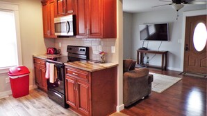 A kitchen with wooden cabinets, a microwave, and a stove. There is a red trash bin and a towel hanging on the oven door. A brown chair is in the living area with a TV on a stand. - Serene, Modern, Cottage, access to a 60 Acre Lake  (Potosi)