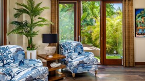 Una habitación con una planta grande en maceta, dos sillones con estampado, una mesita con lámpara y vistas a un follaje exuberante a través de una puerta corrediza de vidrio.