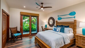 Un dormitorio con cama de madera, ventilador de techo, ventana con vistas a vegetación y una silla junto a la ventana.