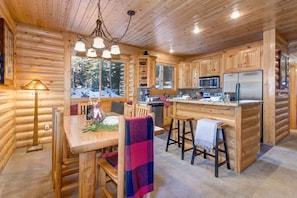 Interior de cabana de tronco com mesa de jantar de madeira, cadeiras, área de cozinha com geladeira, micro-ondas e fogão, e uma janela grande com vista para uma paisagem nevada.