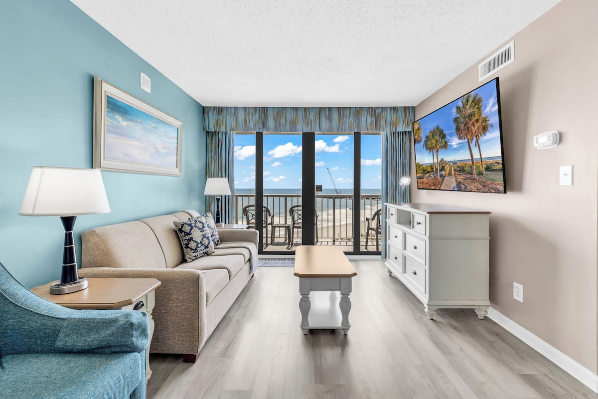 A living room with a sofa, a coffee table, a TV, and a view of the beach through large windows.