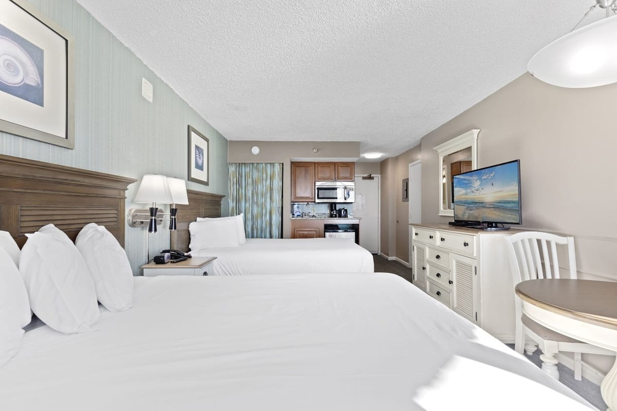A hotel room with two beds, a desk with a chair, a television, a kitchenette, and a painting on the wall.