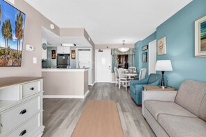 A modern living room with a sofa, a dining table with chairs, a kitchen area, and a hallway leading to a door. - Breakers Boutique Condos | Oceanfront (Myrtle Beach)