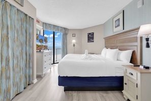 A hotel room with a bed, a nightstand, a lamp, a dresser, and a view of the ocean. - Breakers Boutique Condos | Oceanfront (Myrtle Beach)