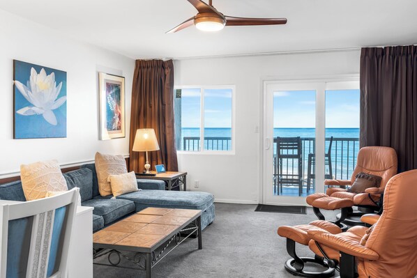 A room with a sofa, a massage chair, a table, a lamp, and a view of the ocean through large windows.