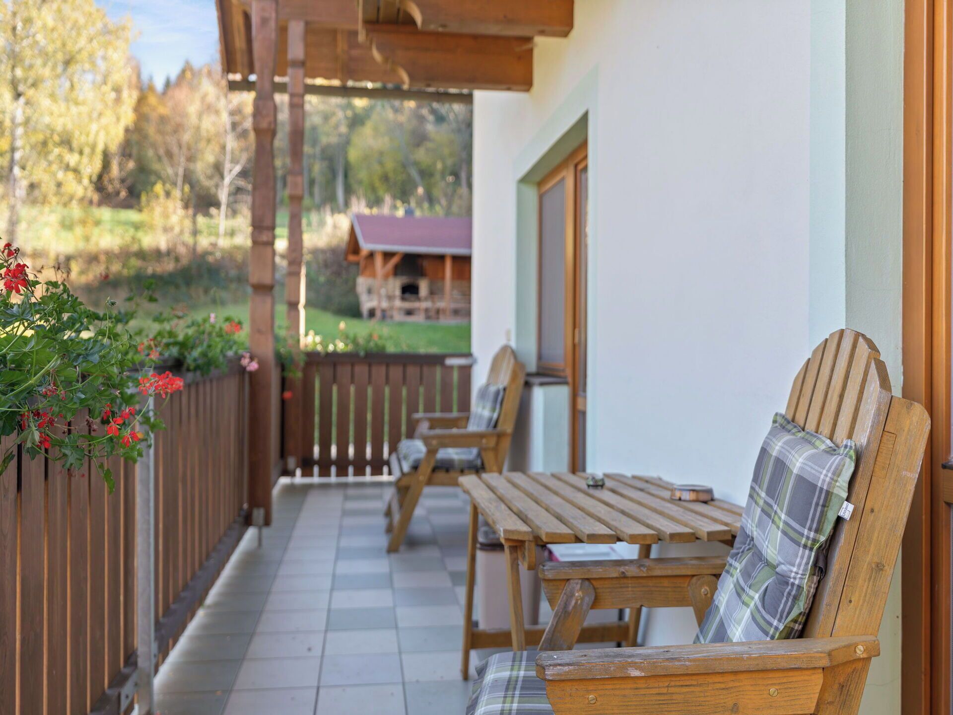 A wooden deck with two wooden chairs and a table, a wooden fence, and a building with a red roof in the background.