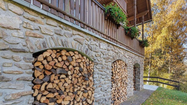 Un edificio de piedra con leña apilada, balcones de madera con macetas de flores y un camino de piedra que conduce a una terraza de madera.