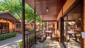 A covered outdoor area with wooden benches and tables, surrounded by wooden cabins with large windows.