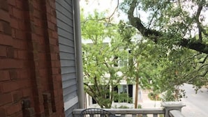 Un balcone con un divano in vimini e un tavolo, affaccia su una strada con alberi e edifici.