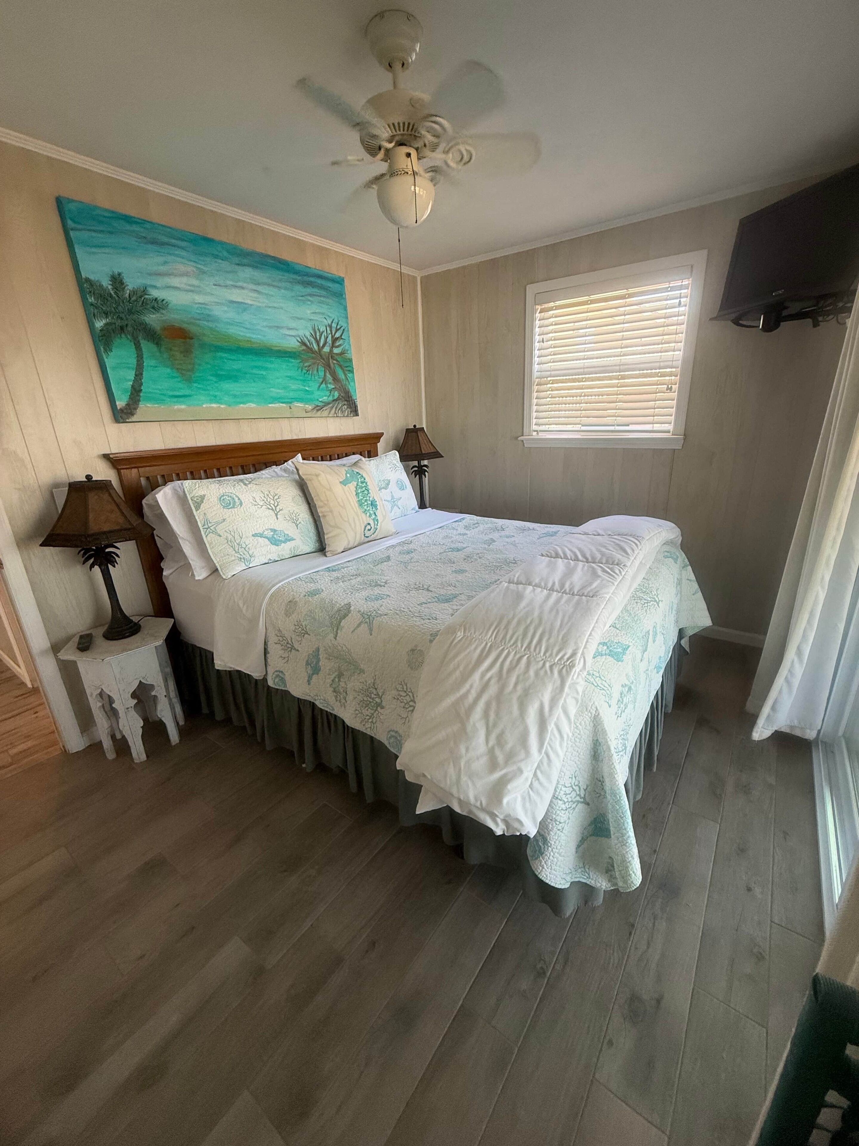 Une chambre à coucher avec un lit, un ventilateur de plafond, un tableau représentant une scène de plage, une fenêtre avec des stores et un téléviseur fixé au mur.