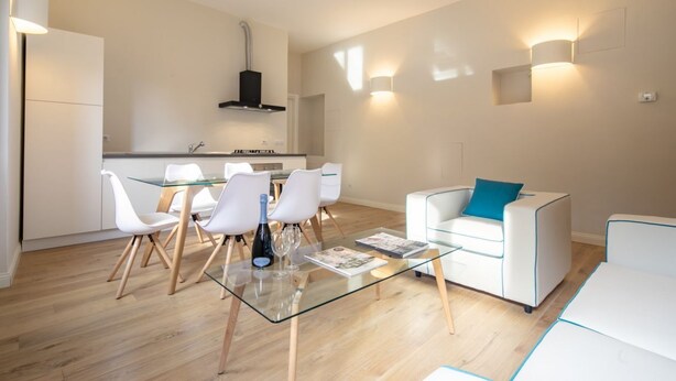 A modern living room with a glass coffee table, white chairs, a white sofa with a blue pillow, and a kitchen area in the background. - Ponte Vecchio White in Florence by Mmega (Firenze)