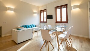 A modern living room with a glass dining table, white chairs, a white sofa with blue cushions, and two wall-mounted lights. - Ponte Vecchio White in Florence by Mmega (Firenze)