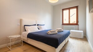 A bedroom with a bed, bedside table, window, radiator, and a ceiling light fixture. - Ponte Vecchio White in Florence by Mmega (Firenze)