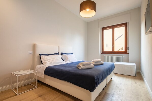 Un dormitorio con cama, mesita de noche, ventana, radiador y lámpara en el techo.