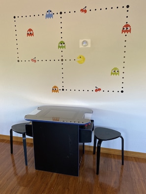 Um quarto com piso de madeira, uma mesa preta com tampo de vidro, duas cadeiras pretas e uma parede decorada com um labirinto do Pac-Man.