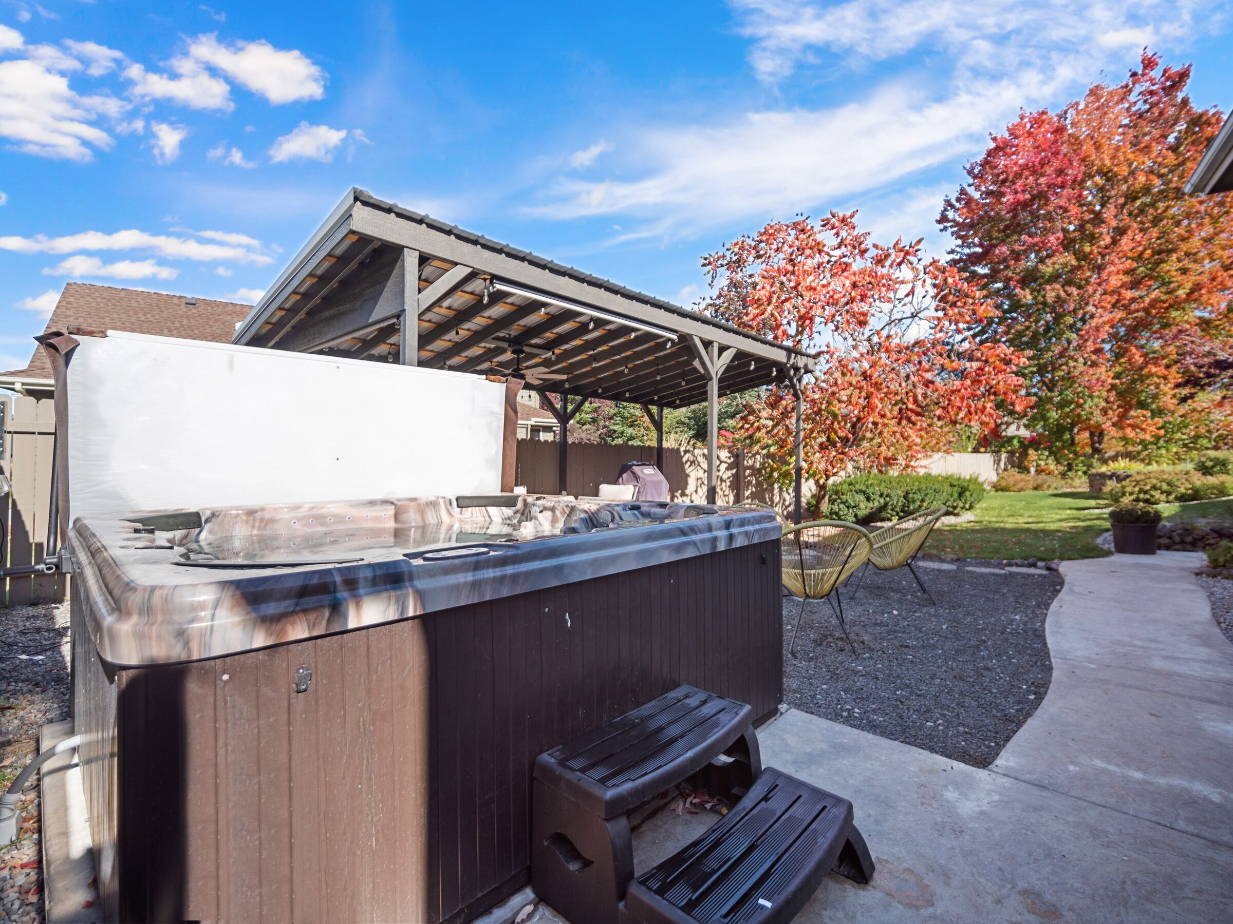 A hot tub with a wooden exterior, a covered seating area, and a patio with a tree displaying autumn foliage.