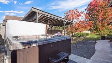 A hot tub with a wooden exterior, a covered seating area, and a patio with a tree displaying autumn foliage.