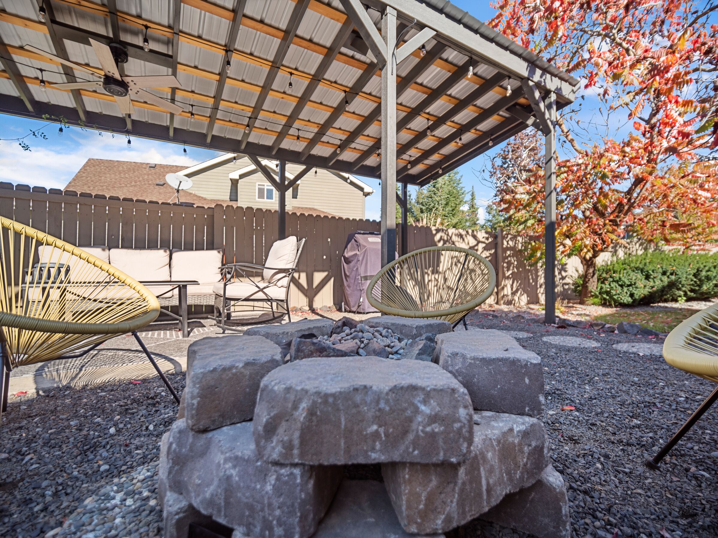 A covered patio area with a fire pit, wicker chairs, and a wooden table.