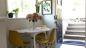 Um espaço de refeições pequeno com uma mesa, cadeiras amarelas e um vaso de flores. Há uma janela com plantas no parapeito e uma porta que dá para uma escadaria.