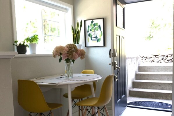 A small dining area with a table, yellow chairs, and a vase of flowers. There is a window with plants on the sill and a door leading to a staircase.