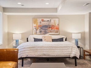 A bedroom with a bed, two bedside tables, a chair, and a large abstract painting on the wall.