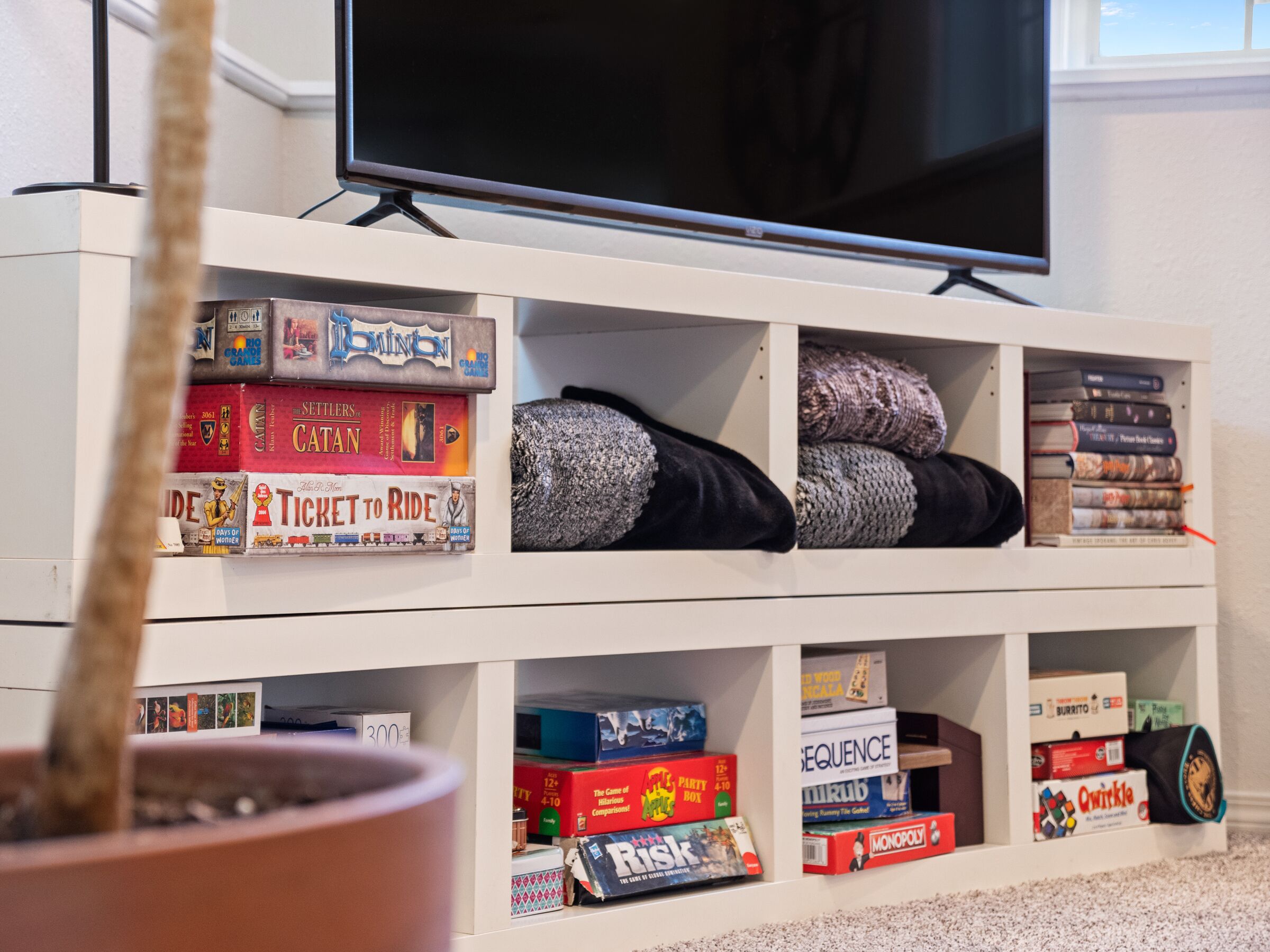 A room with a TV mounted on the wall, a bookshelf filled with board games, and a potted plant.