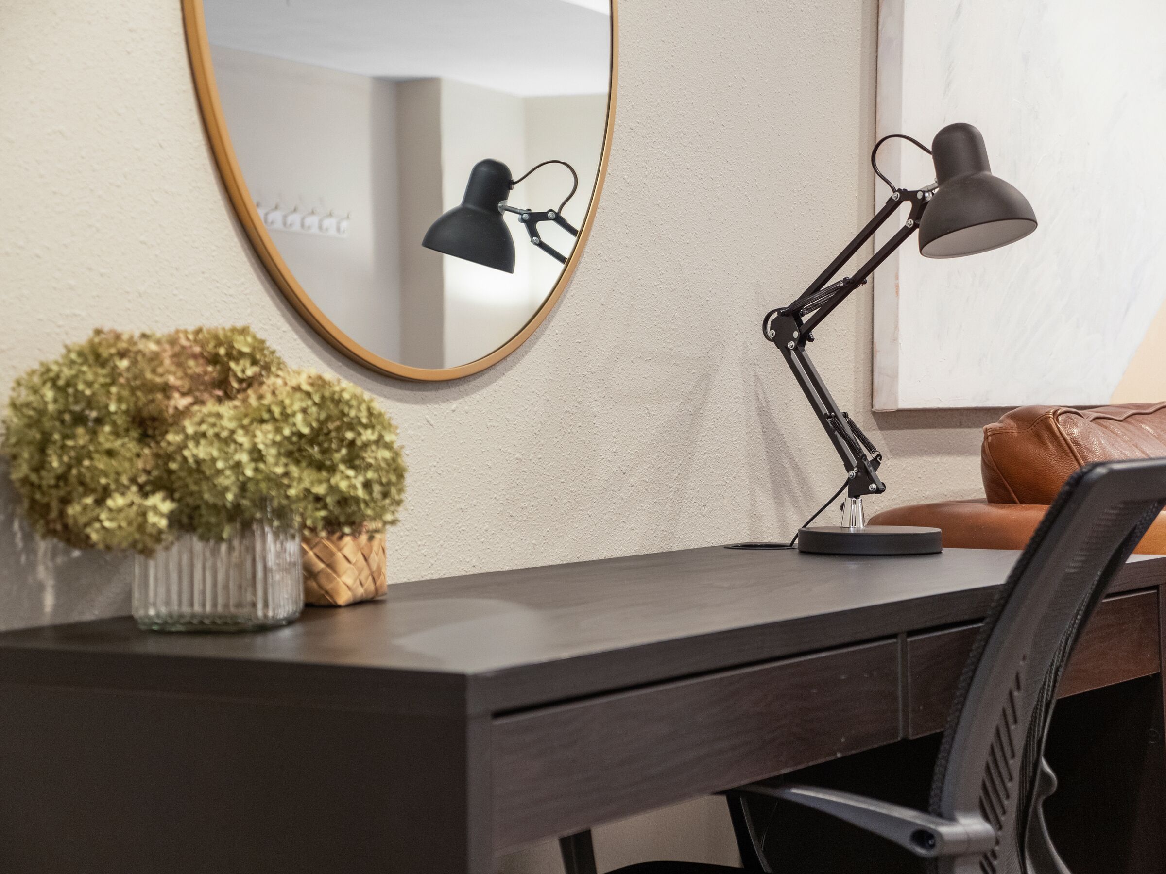 A desk with a dark surface, a round mirror with a gold frame, two black desk lamps, a small potted plant, and a brown leather chair.