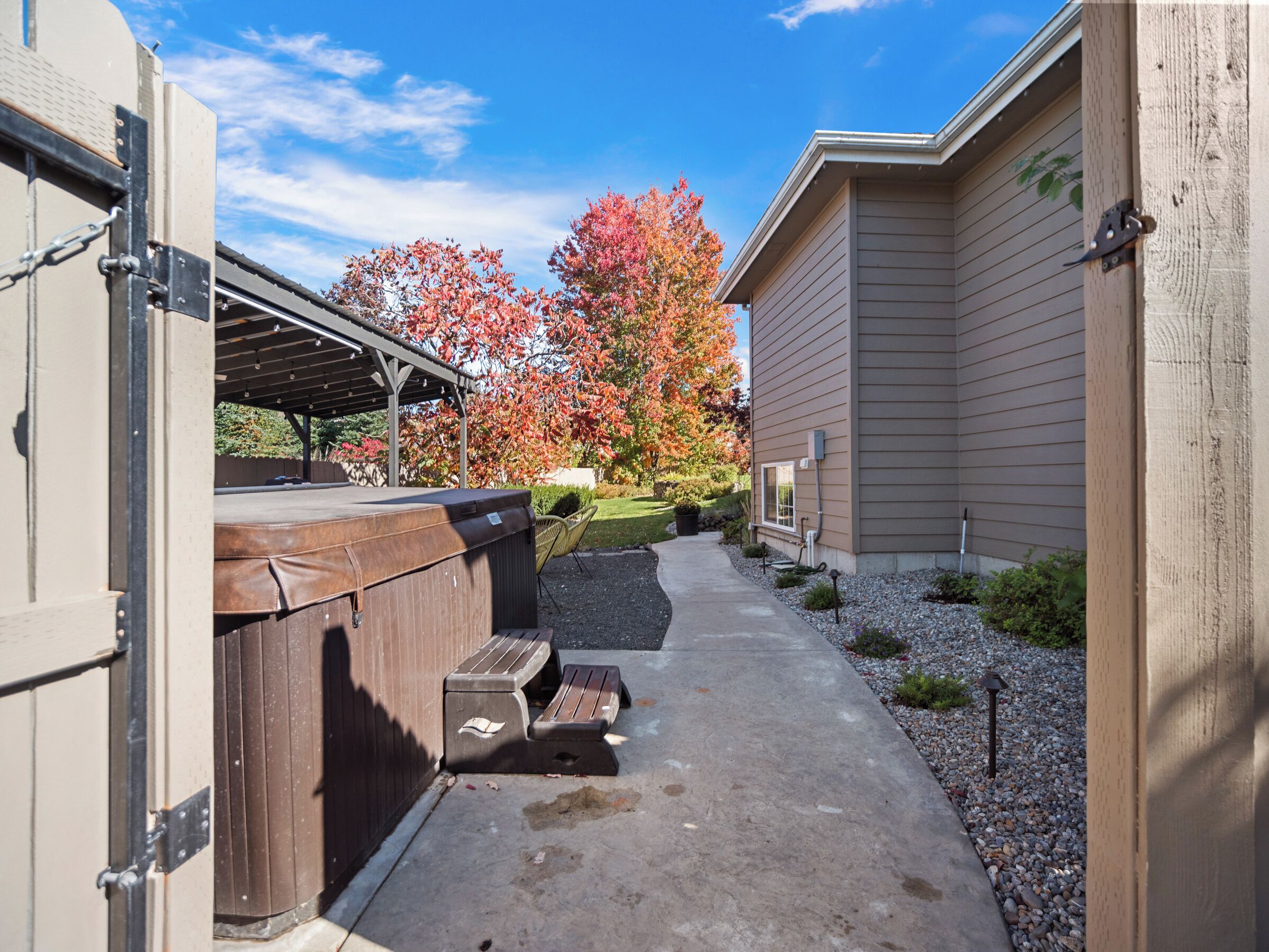 A covered outdoor area with a hot tub, a concrete pathway, and a building with siding.