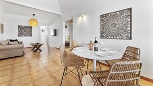 Una sala de estar moderna con una mesa de comedor redonda, sillas de mimbre y un sofá. Se observan elementos decorativos en las paredes y un candelabro en el techo.