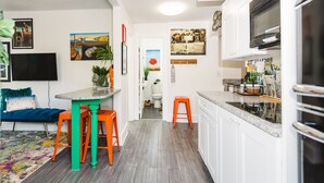 Una cocina moderna con armarios blancos, encimera de granito y horno integrado. Cuenta con una zona de comedor con mesa y sillas, y un televisor montado en la pared.