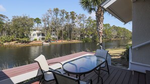 A deck with a glass table and chairs overlooking a lake, surrounded by trees and a building.