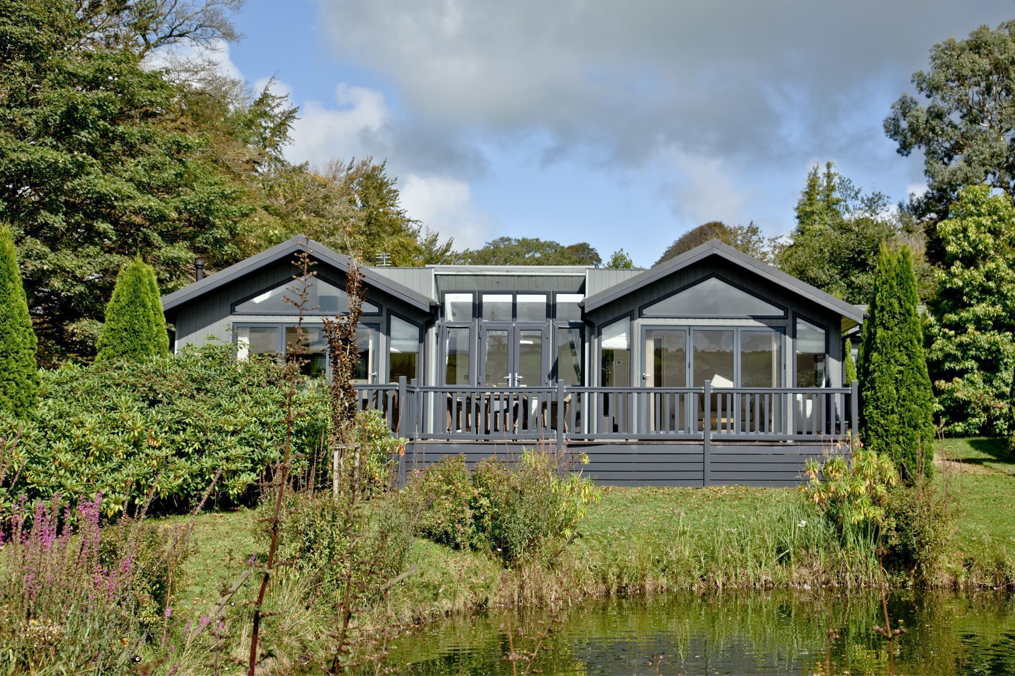 A modern two-story lodge with large windows and a deck, surrounded by lush greenery and a pond.