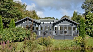 A modern two-story lodge with large windows and a deck, surrounded by lush greenery and a pond. - Watermouth Lodge, Kentisbury Grange (Barnstaple)