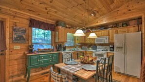 A wooden cabin interior with a dining table set for four, kitchen cabinets, a refrigerator, and a window with blinds.