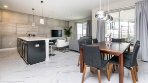 A modern kitchen with a central island, a dining area with a wooden table and chairs, and a living area with a sofa and a TV.