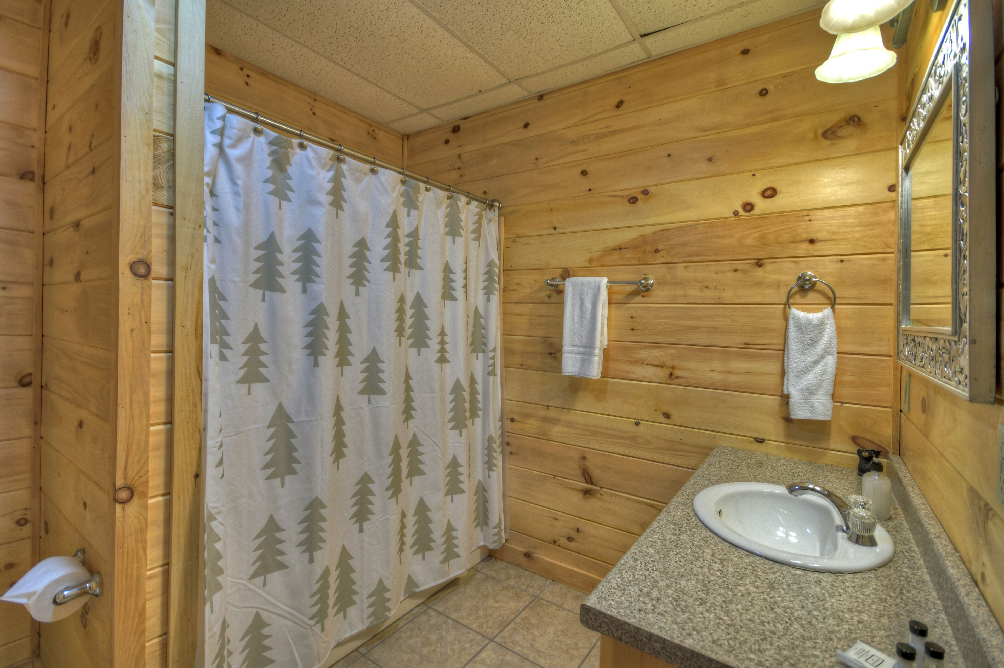 Une salle de bain rustique, avec des murs en bois, comprenant une douche avec un rideau de douche orné d’un motif d’arbre, un lavabo avec un robinet et un porte-serviettes avec deux serviettes.